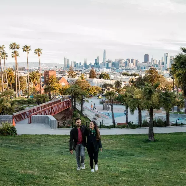 Una pareja camina hacia la cámara con Dolores Park y el Skyline de 贝博体彩app detrás de ellos.